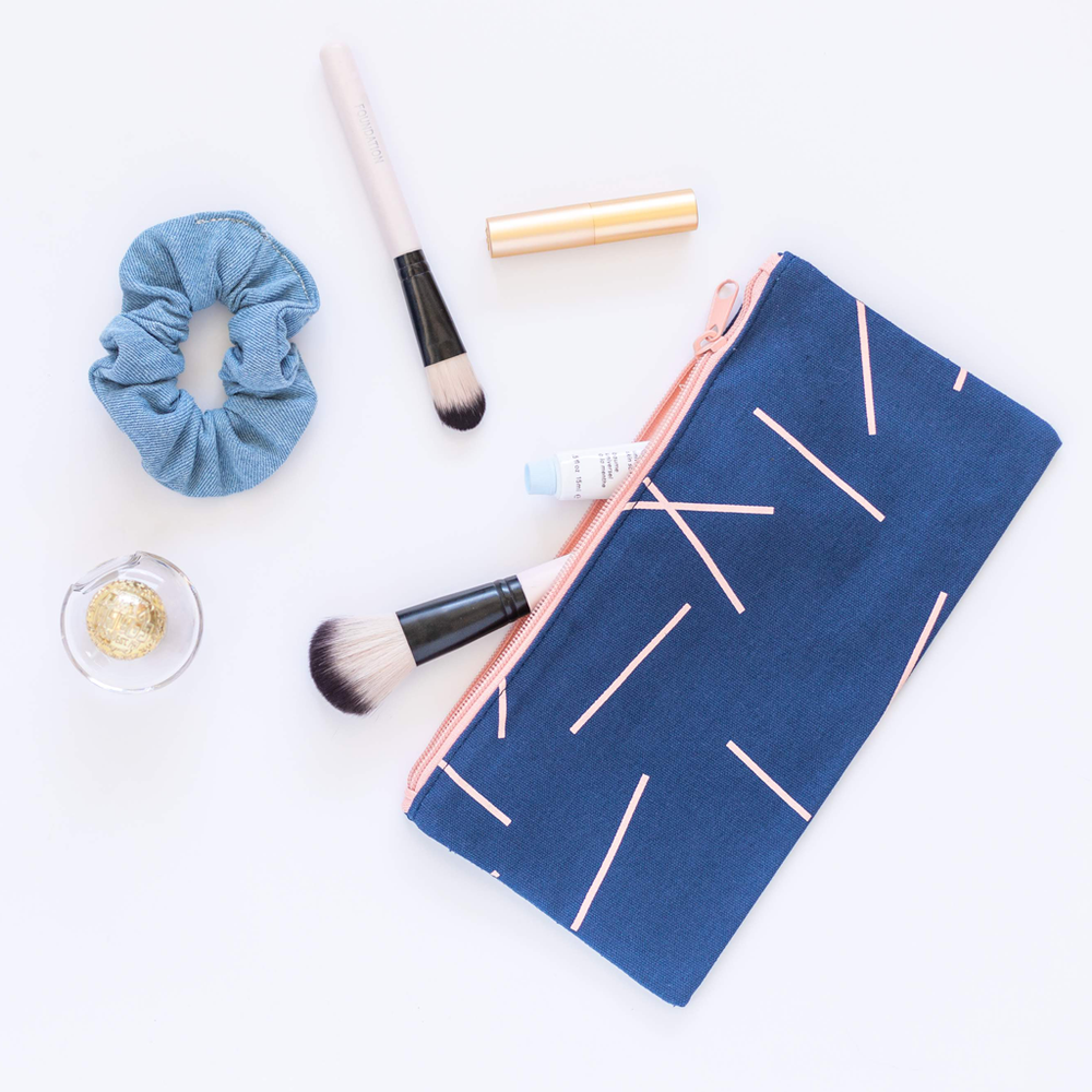 Blue makeup pouch with geometric pattern, blue scrunchie, makeup brushes, and small bottle on a white background