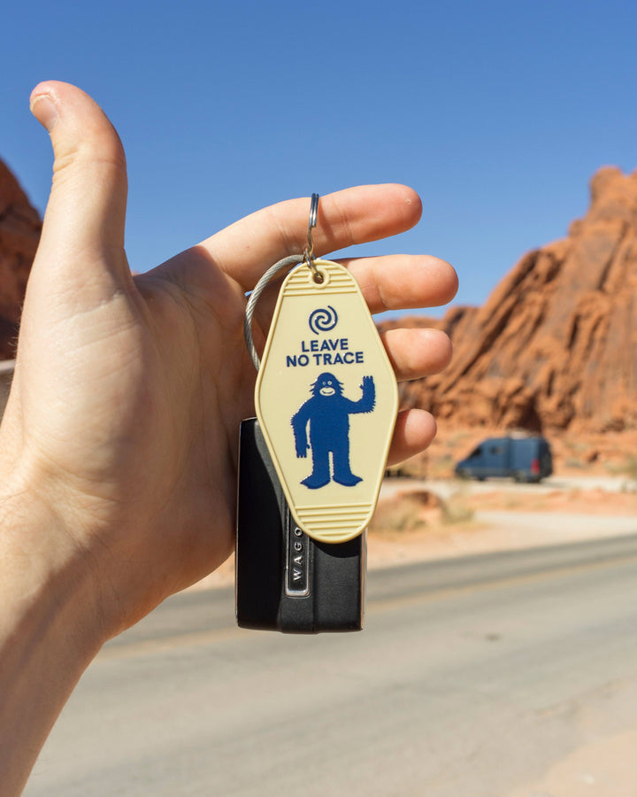 Hand holding a keychain with 'Leave No Trace' logo against a desert landscape.