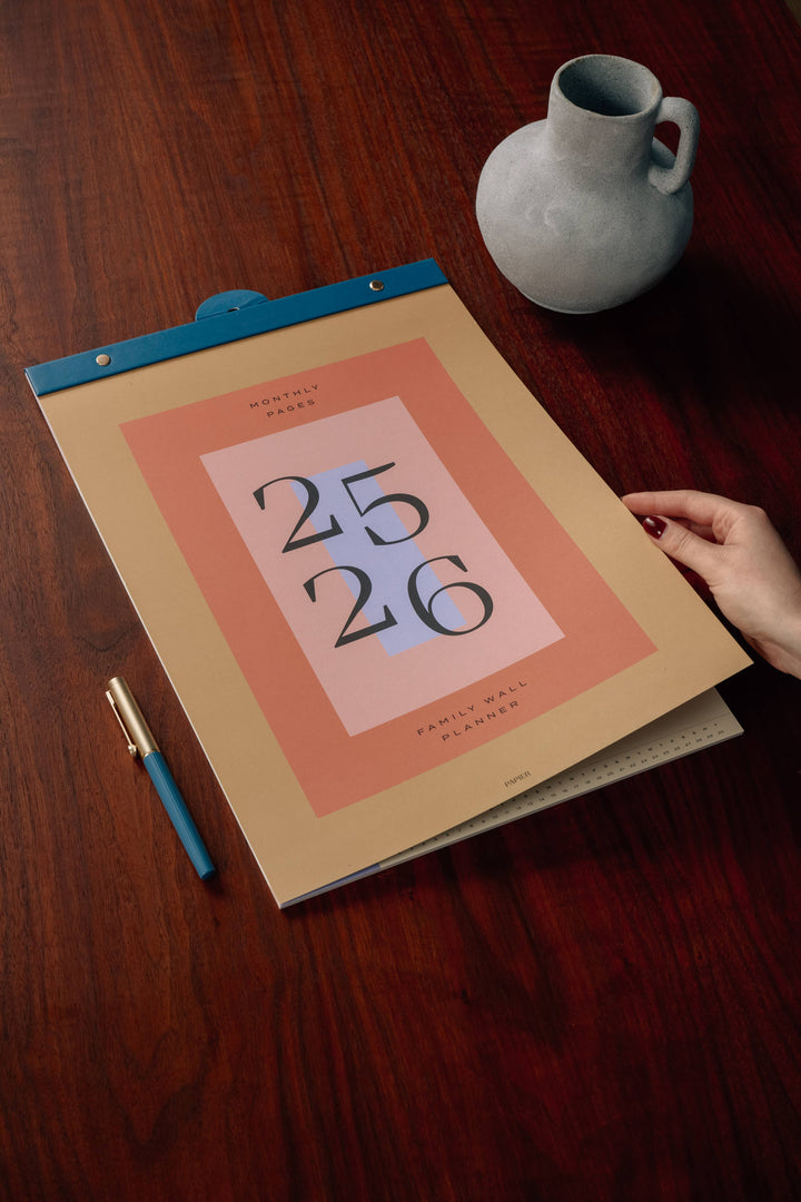 Hand holding a planner with '25 26' on a wooden surface with a vase and pen.