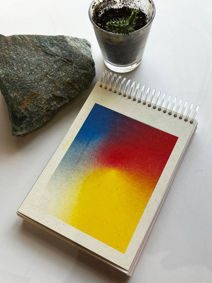 Notebook with a colorful cover next to a rock and a glass of dark liquid on a light surface