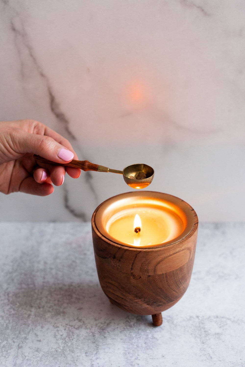 Wooden container with a lit candle and a hand holding a  wax spoon over it on a marble surface.