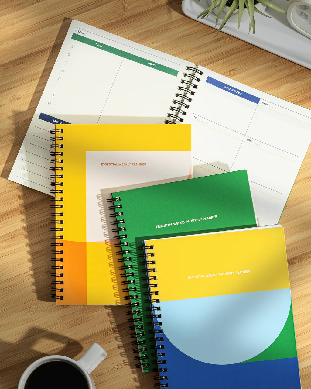 Colorful weekly planners on a wooden surface with a coffee cup and plant in the background.