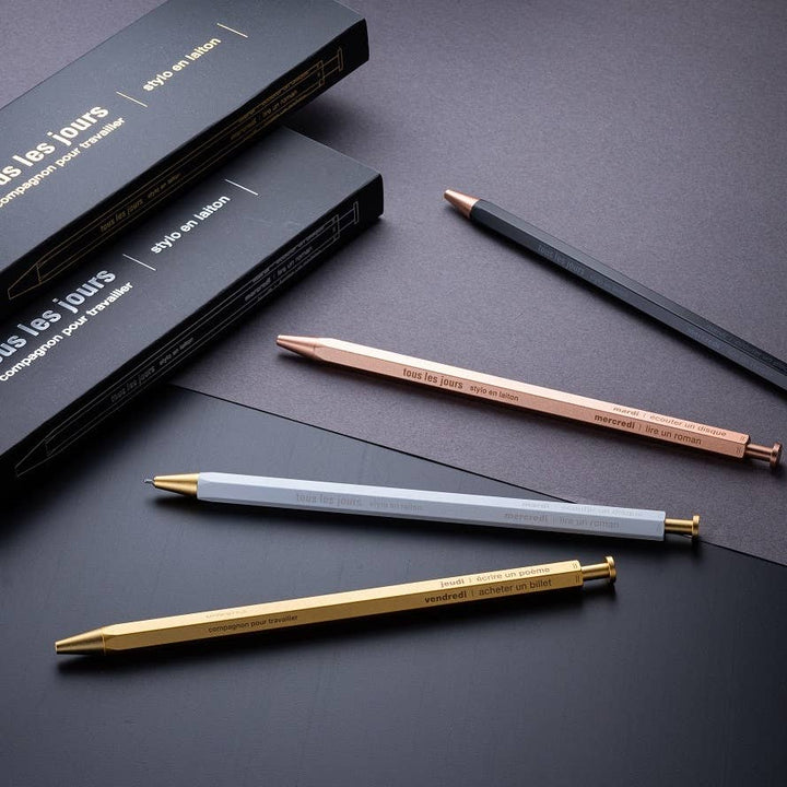 A selection of ballpoint pens with brass bodies, available in gold, silver, and rose gold colors, displayed alongside their packaging boxes.