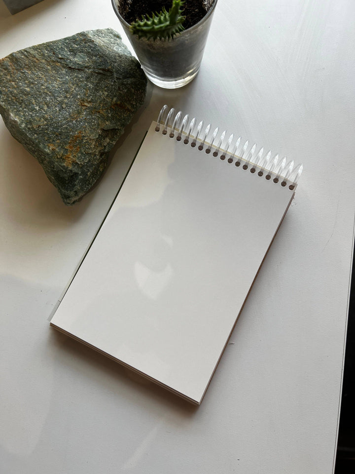Blank spiral-bound notebook on a white surface with a rock and glass container in the background.