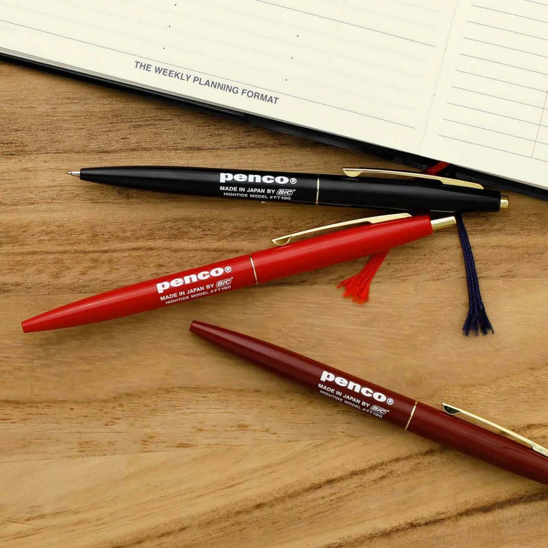 Three pens with 'penco' branding on a wooden surface next to a planner.